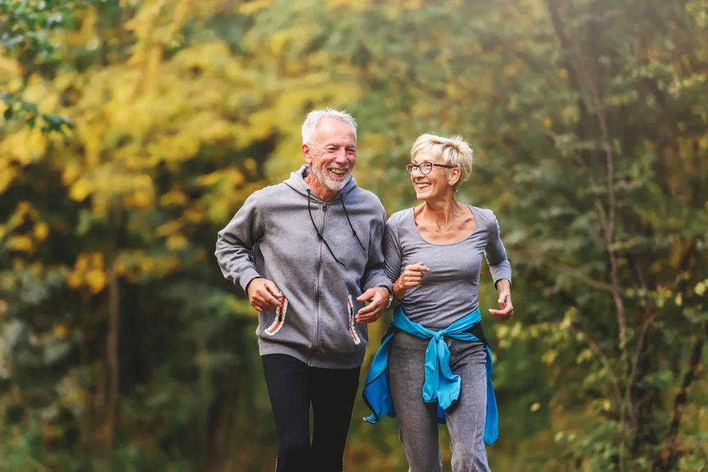 An older adult couple, man and wide, smiling while jogging outside together in a beautiful park with trees in background, symbolizing new advancements in Hearing Aid Health Monitoring Features